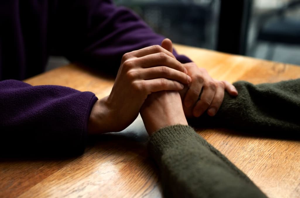 Two people holding hands in comfort over a wooden table