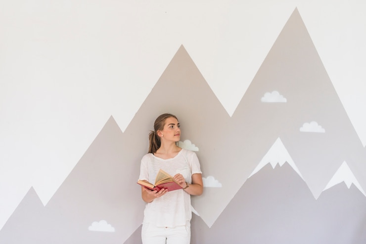 a young woman stands with a book against wallpaper with mountains in the background