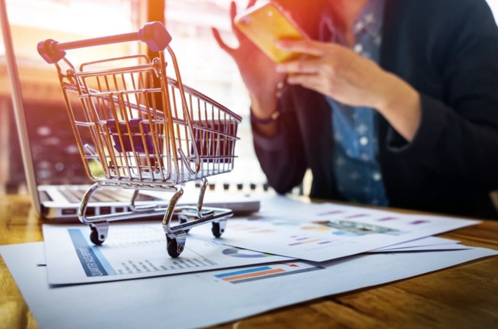 A shopping cart on top of a wooden desk with financial charts, a person in the background